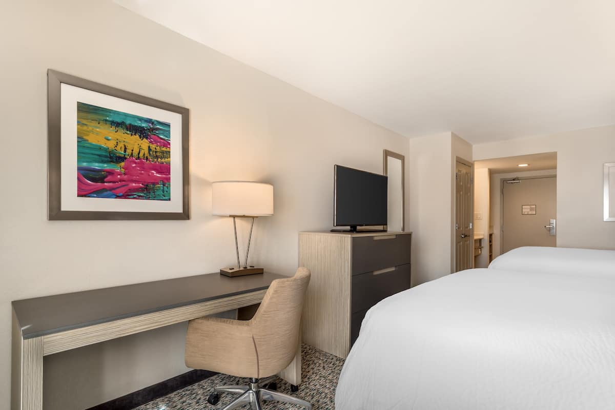 A hotel room with a bed, a desk with a chair, a television, and a painting on the wall.