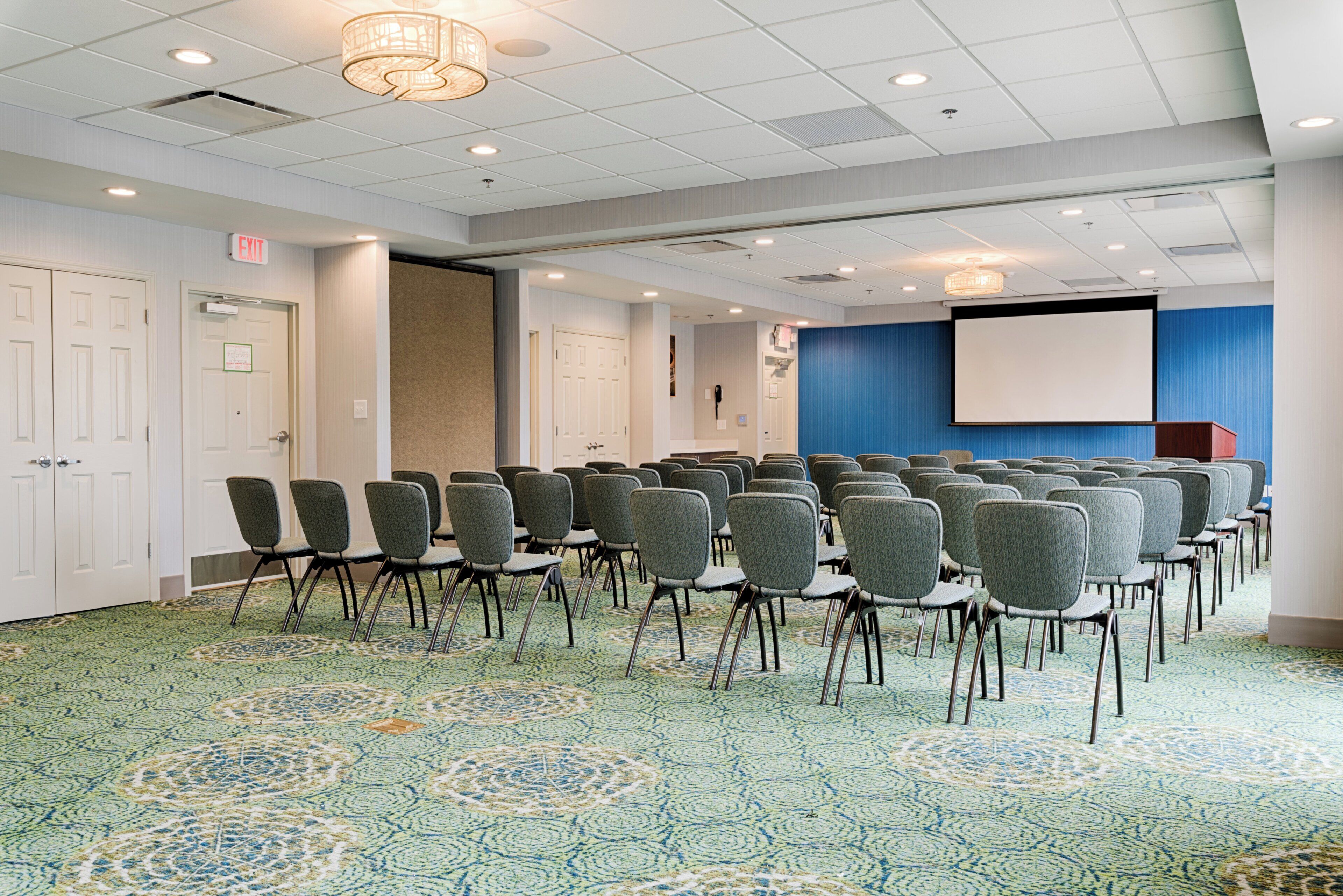A conference room with rows of chairs, a projector screen, and an exit sign.