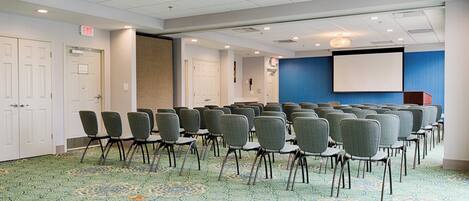 A conference room with rows of chairs, a projector screen, and an exit sign.