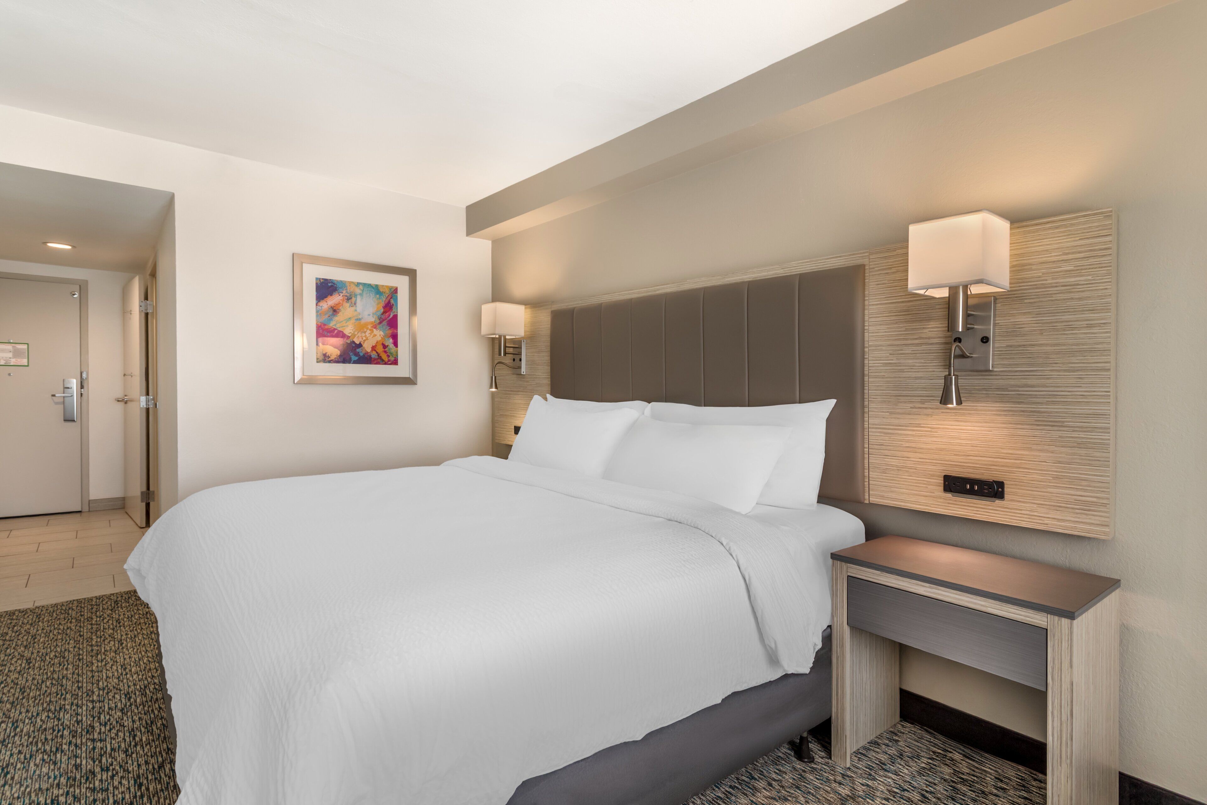 A hotel room with a large bed, two bedside lamps, a headboard, a small desk, and a painting on the wall.