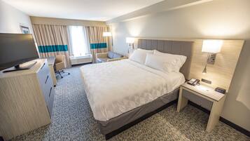 A hotel room with a large bed, a desk with a computer, a chair, a TV, and a bedside table with a lamp and phone.
