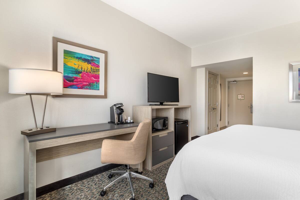 A hotel room with a bed, a desk with a television, a chair, and a painting on the wall.