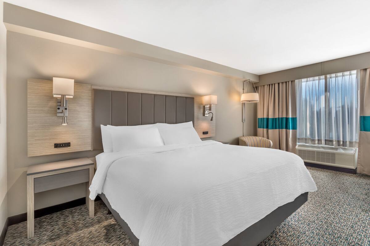 A hotel room with a large bed, two bedside tables, a wall-mounted lamp, a window with curtains, and a built-in air conditioning unit.