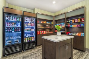 A kitchenette with two refrigerators stocked with beverages and snacks, a small island with a plant, and cabinets with packaged food items. - Holiday Inn Knoxville N - Merchant Drive by IHG (Knoxville)