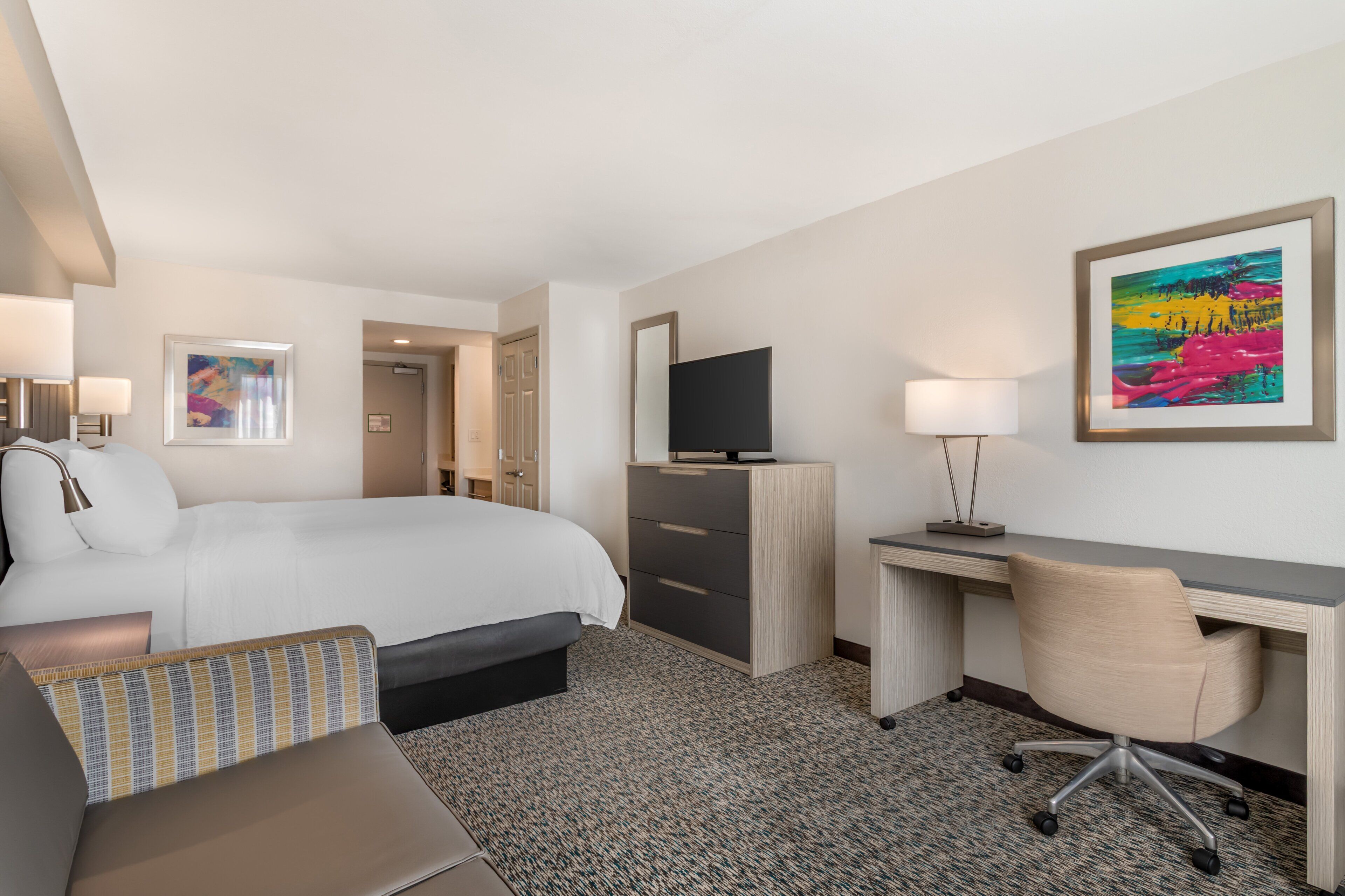 A hotel room with a bed, desk, chair, TV, and artwork.