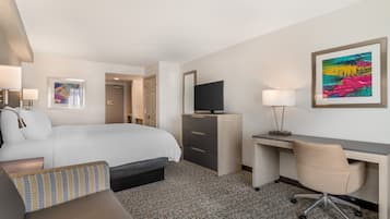 A hotel room with a bed, desk, chair, TV, and artwork.