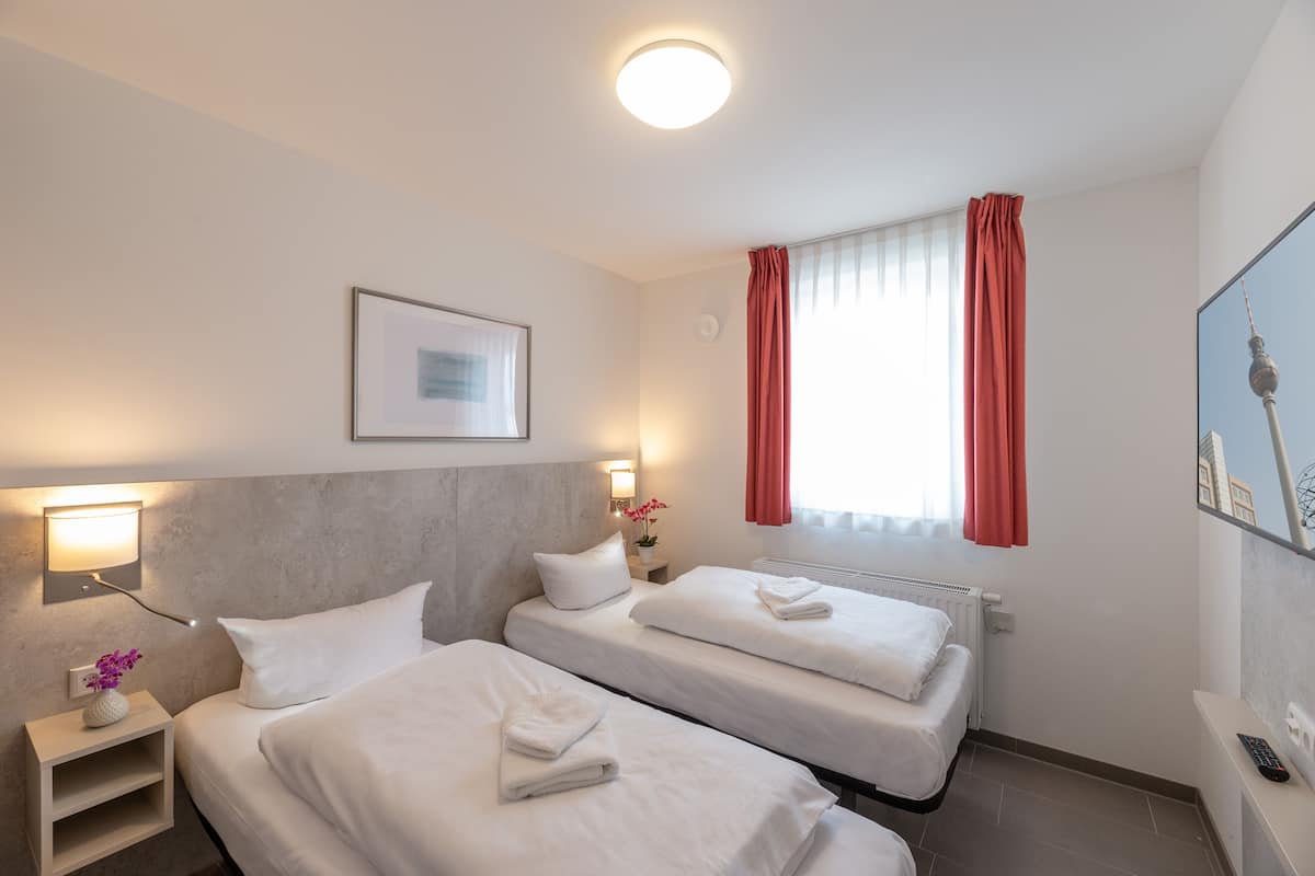 Habitación de hotel con dos camas, una mesita de noche con lámpara, un cuadro en la pared y una ventana con cortinas.