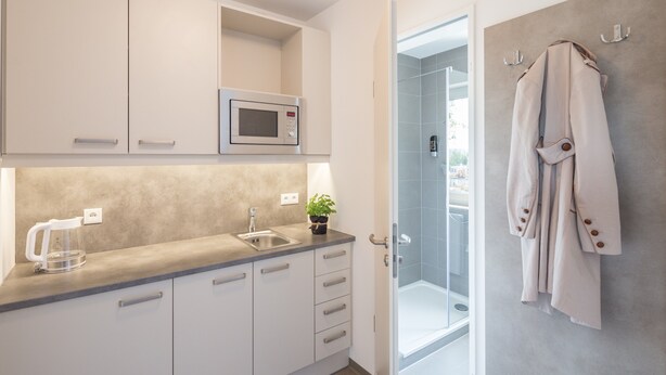 A modern kitchen with white cabinets, a microwave, a kettle, and a sink. There is a glass shower door leading to a bathroom with a towel rack. - TRIO Apartment Hotel Berlin (Berlin)