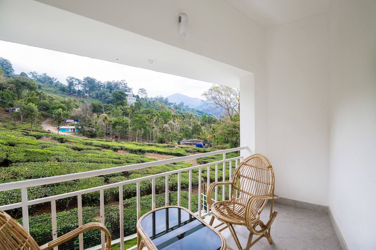 A balcony with wicker chairs and a glass table, overlooking a lush green landscape with tea plantations and distant mountains.