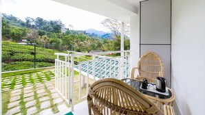 Un balcón con una silla y una mesa de mimbre, barandal blanco y vistas a un paisaje verde exuberante con colinas y un edificio a lo lejos.