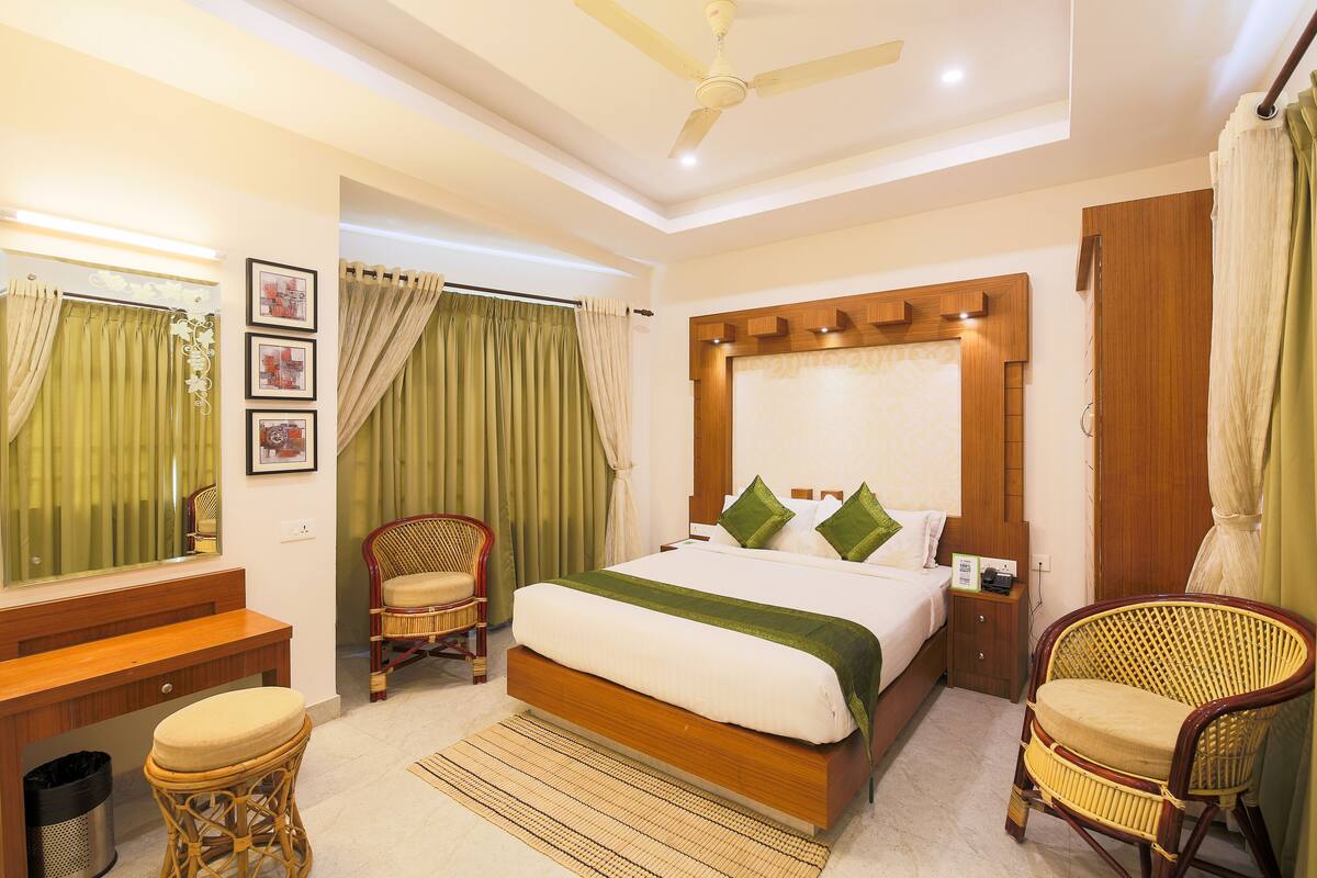 A bedroom with a bed, two wicker chairs, a wooden bedside table, a mirror, and a ceiling fan.