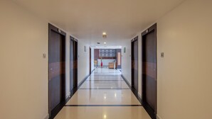 Un pasillo de hotel con diseño moderno, que cuenta con una zona de estar central con un jarrón y una silla, y un papel tapiz con estampado en el fondo.