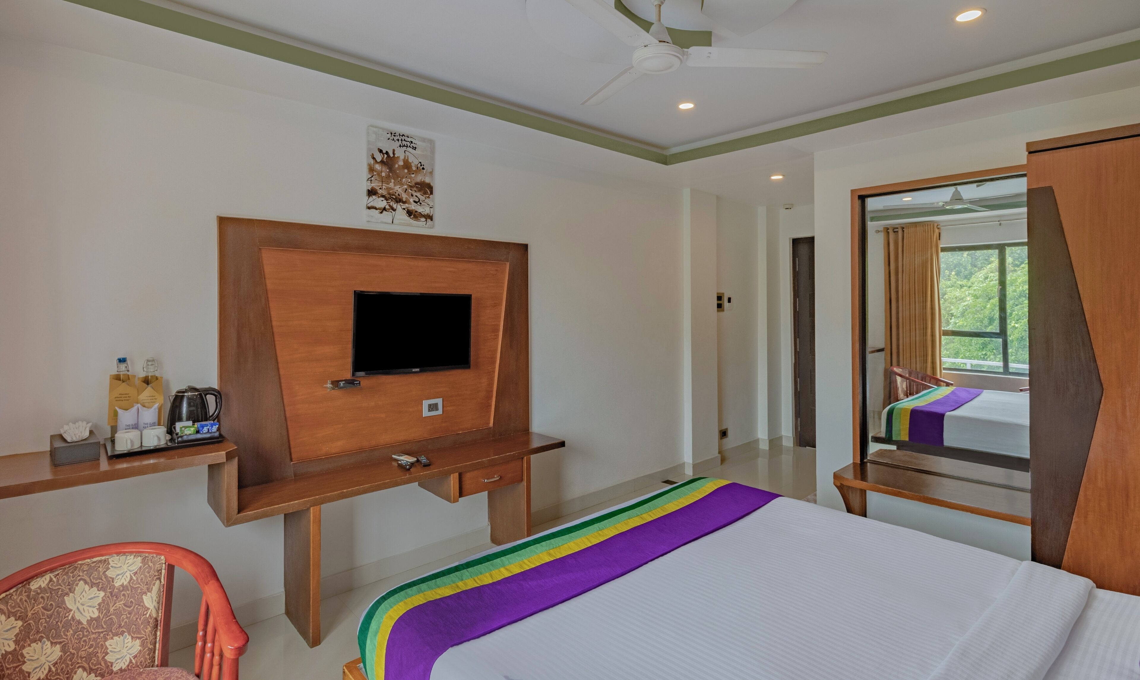 A hotel room with a bed, a TV mounted on a wooden cabinet, a small desk with a kettle and cups, and a mirror reflecting the room.