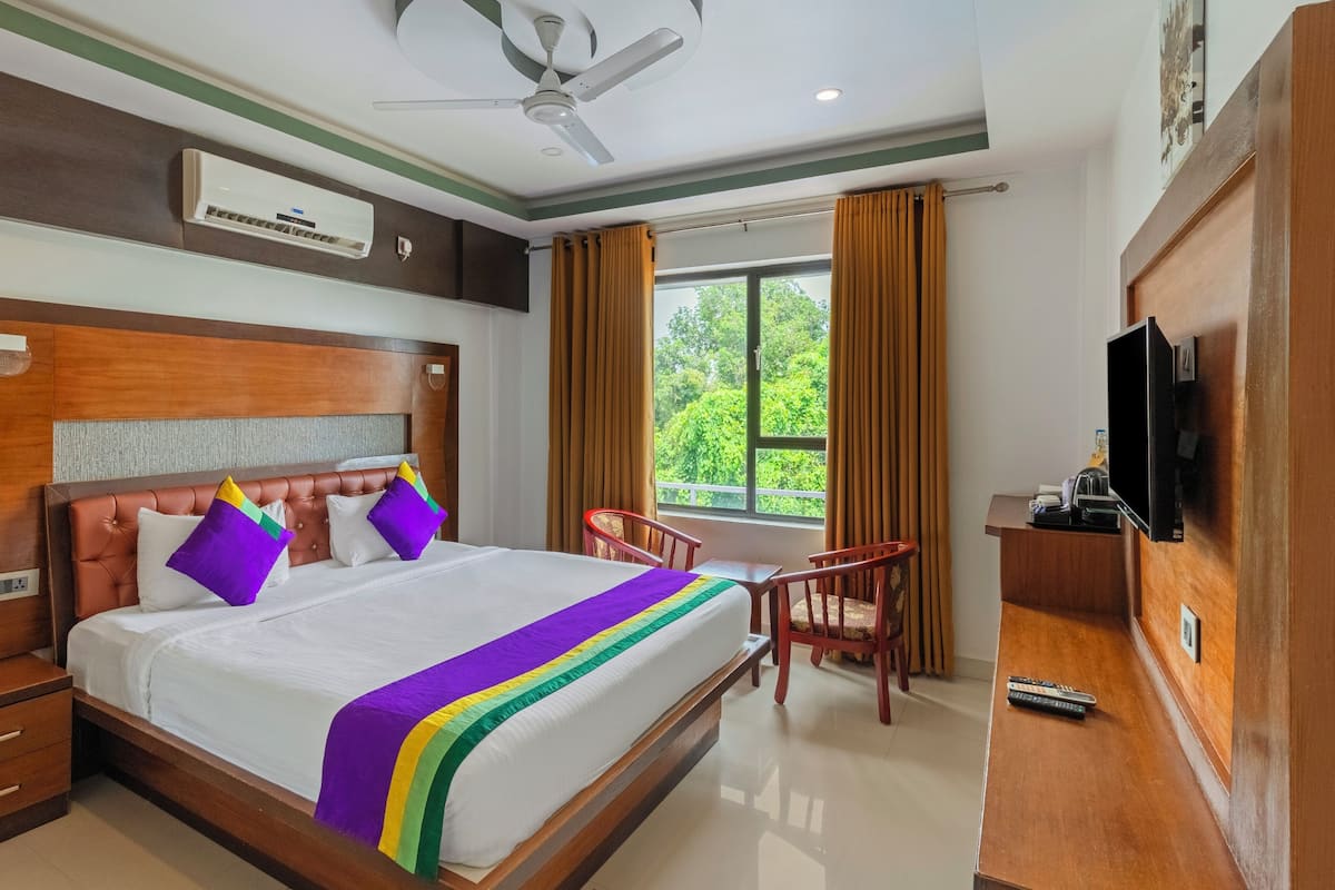 Habitación de hotel con una cama grande, cabecera de madera, ventilador de techo, aire acondicionado, televisor, escritorio con silla y ventana con cortinas.