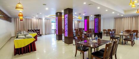 A spacious dining area with multiple tables and chairs, a buffet setup on the left, and decorative lighting.