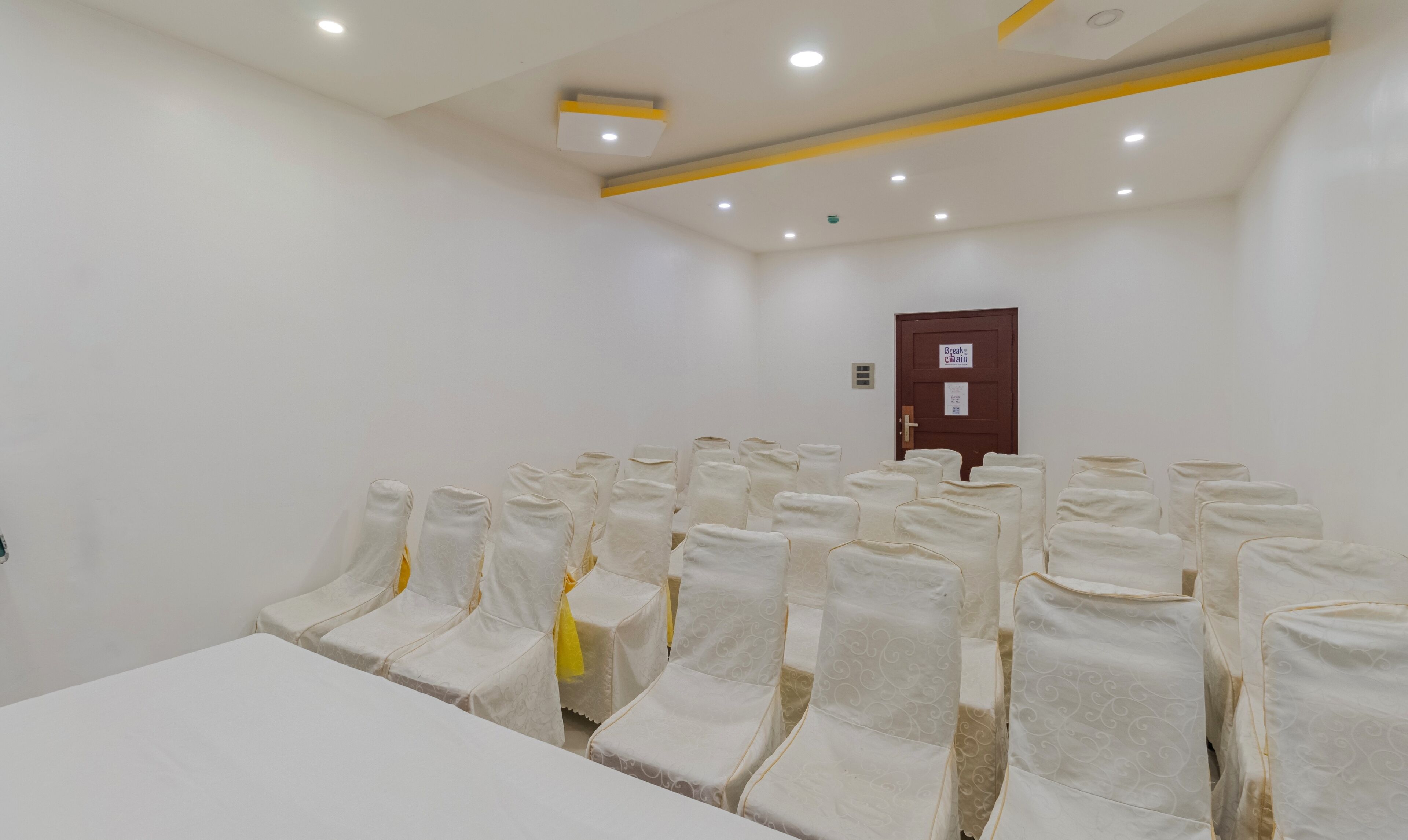 Un salón de conferencias con filas de sillas blancas, una puerta de madera con un letrero y luces empotradas en el techo.