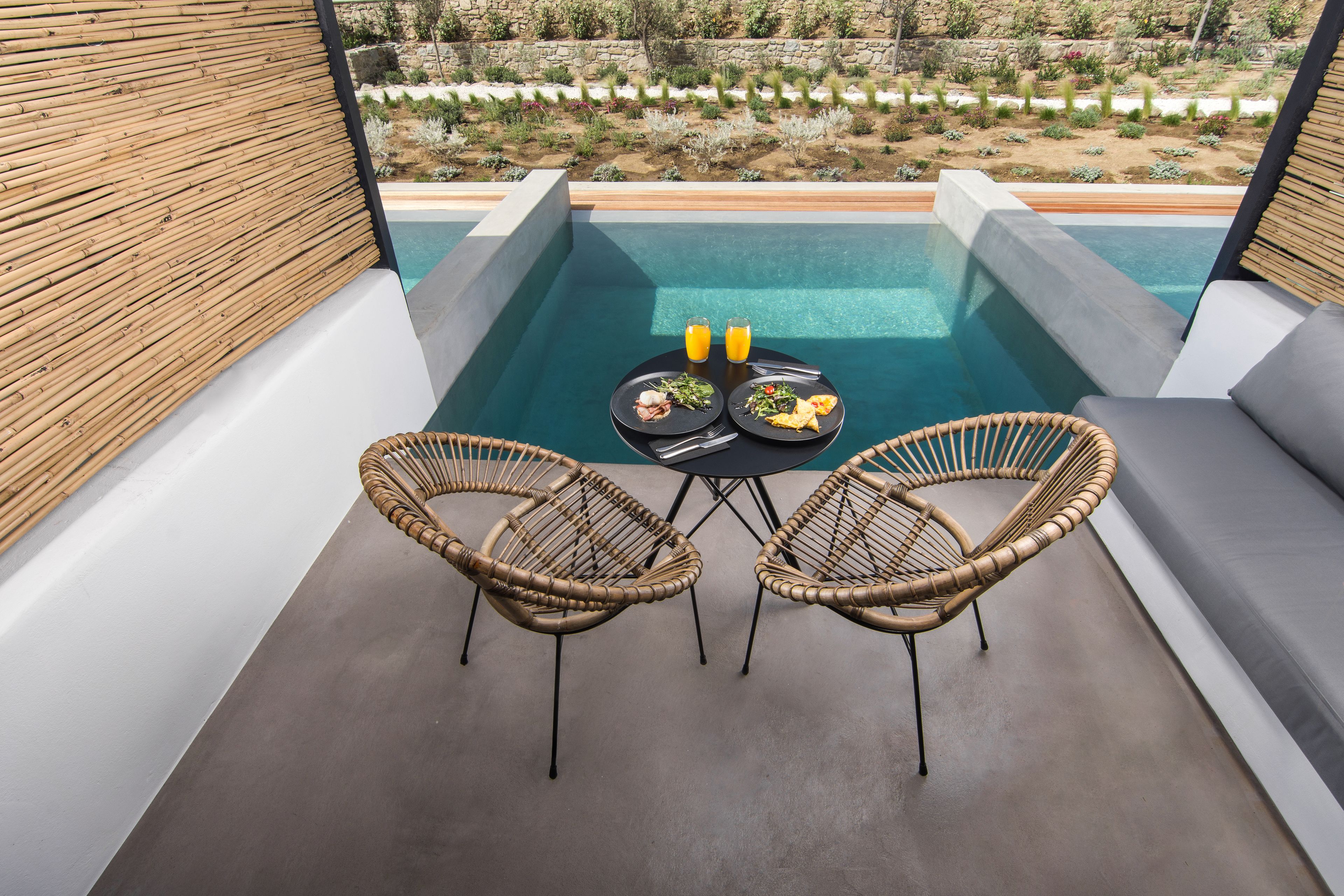 Un espace aménagé au bord de la piscine, avec deux fauteuils en osier, une table sur laquelle sont disposés des assiettes de nourriture et des boissons, et une vue sur un jardin paysager.
