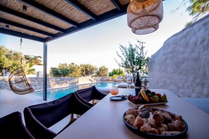 A dining table with a fruit bowl, bread, and wine glasses, overlooking a swimming pool and outdoor seating area. - My Mykonos Boutique by Mr and Mrs White (Mykonos)