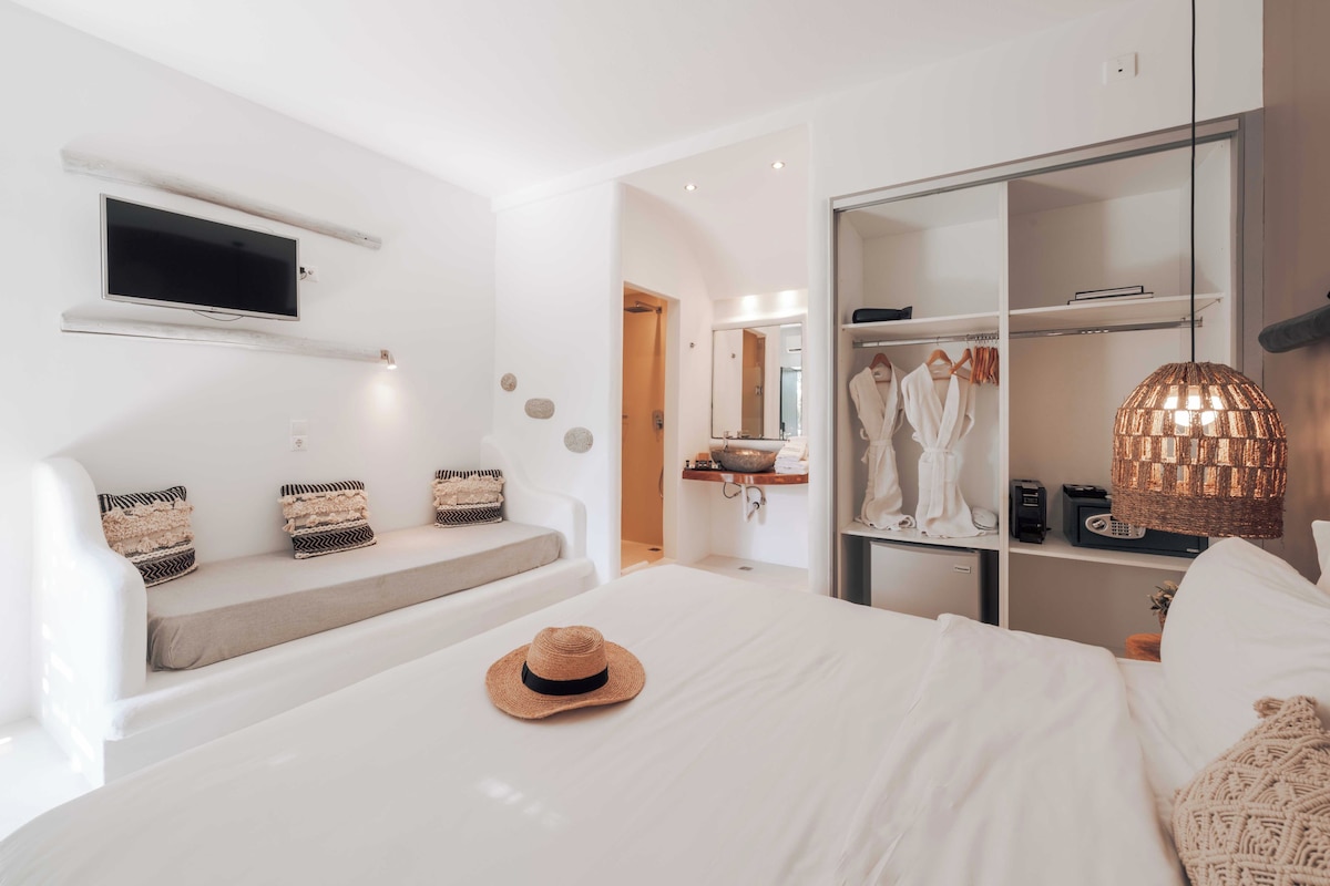 A modern hotel room with a large bed, a TV mounted on the wall, a wardrobe, and a seating area with cushions.