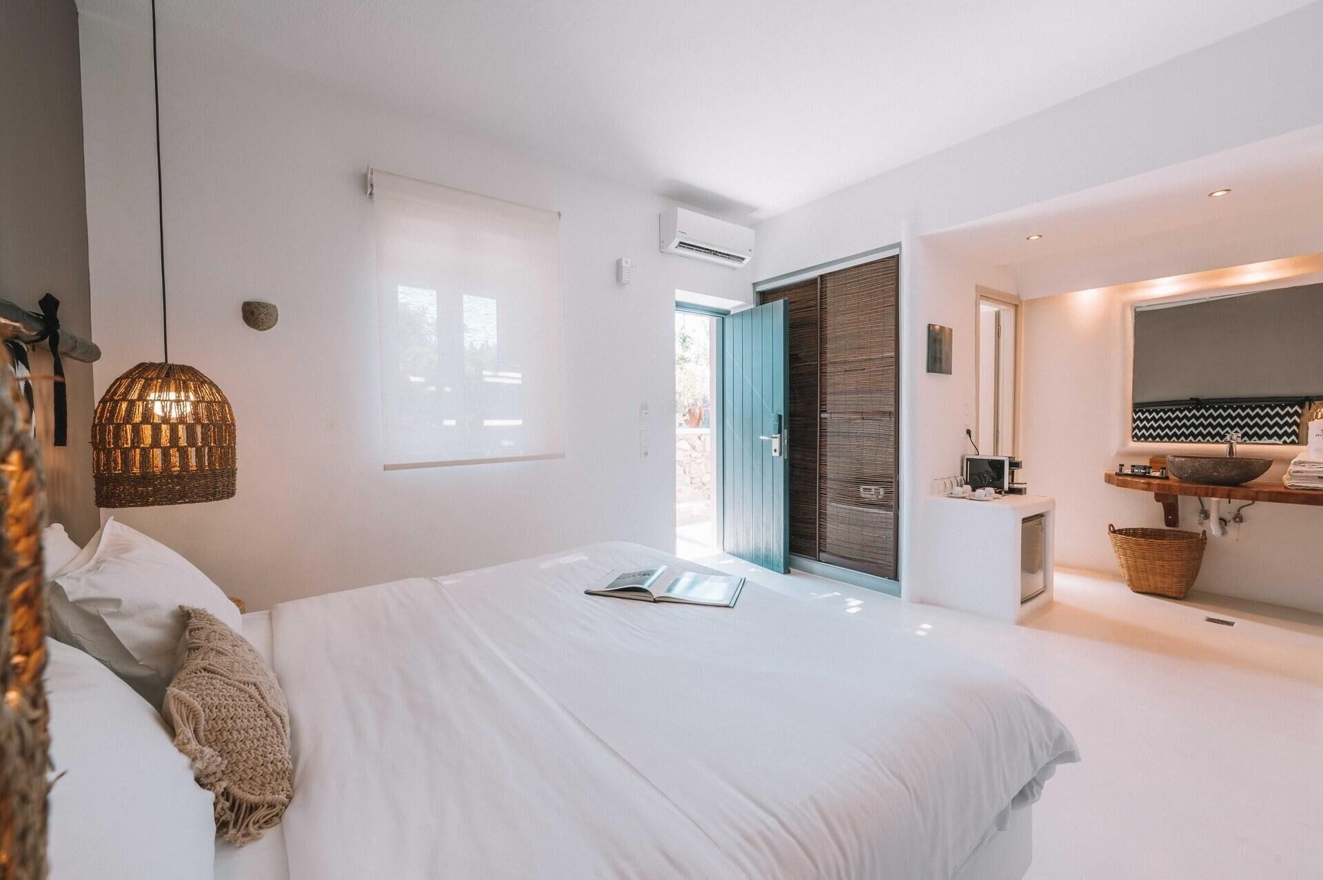Ein modernes Schlafzimmer mit einem großen Bett, einer Pendelleuchte, einem Fenster und einem Badezimmer mit Waschbecken und Spiegel.