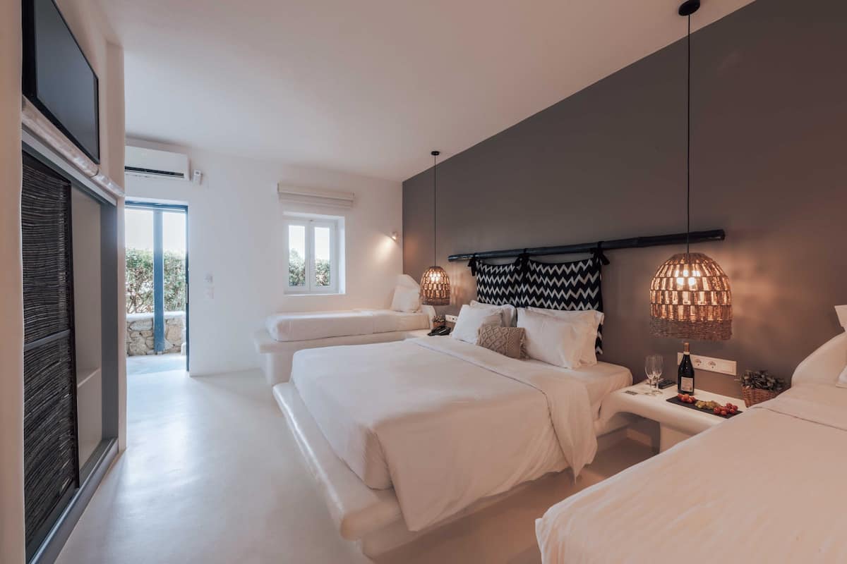 Une chambre d’hôtel moderne avec un grand lit, un lit plus petit et un canapé. On y trouve un téléviseur fixé au mur, une fenêtre donnant sur l’extérieur et un lustre.