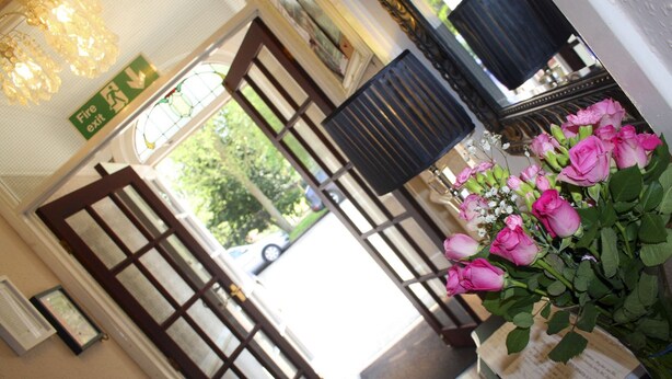 A hotel lobby with a fire exit sign, a large window with a view of the outdoors, and a bouquet of pink roses on a table. - The Sherwood (Skegness)