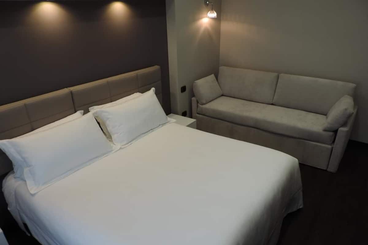 A neatly made bed with white linens, a grey headboard, and a grey sofa in a modern room with a dark wall and a wall-mounted light fixture.