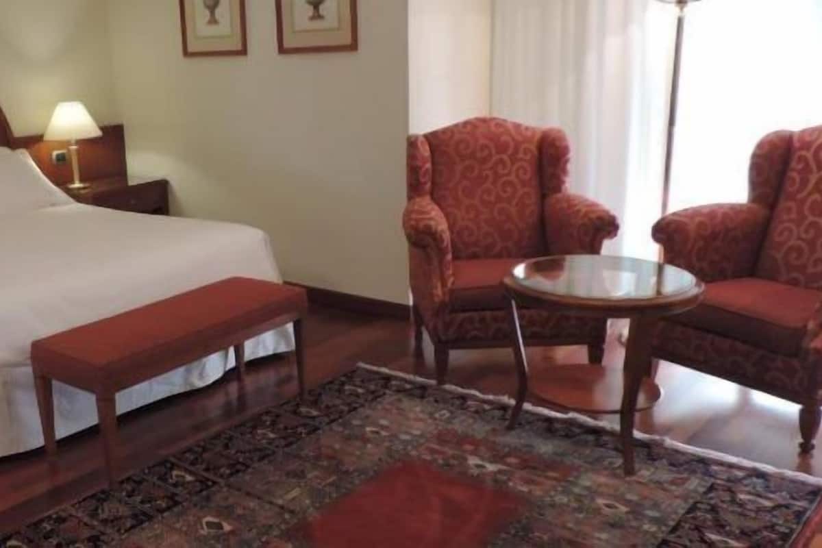 A hotel room with a large bed, a bench, two armchairs, a small table, and a patterned rug.