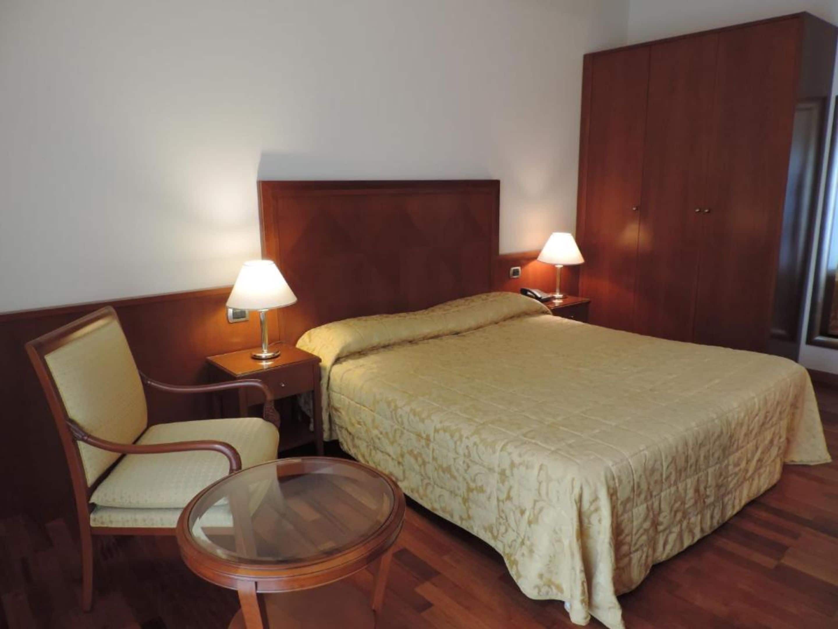 A hotel room with a large bed, a wooden wardrobe, a chair, a small table, and two bedside lamps.