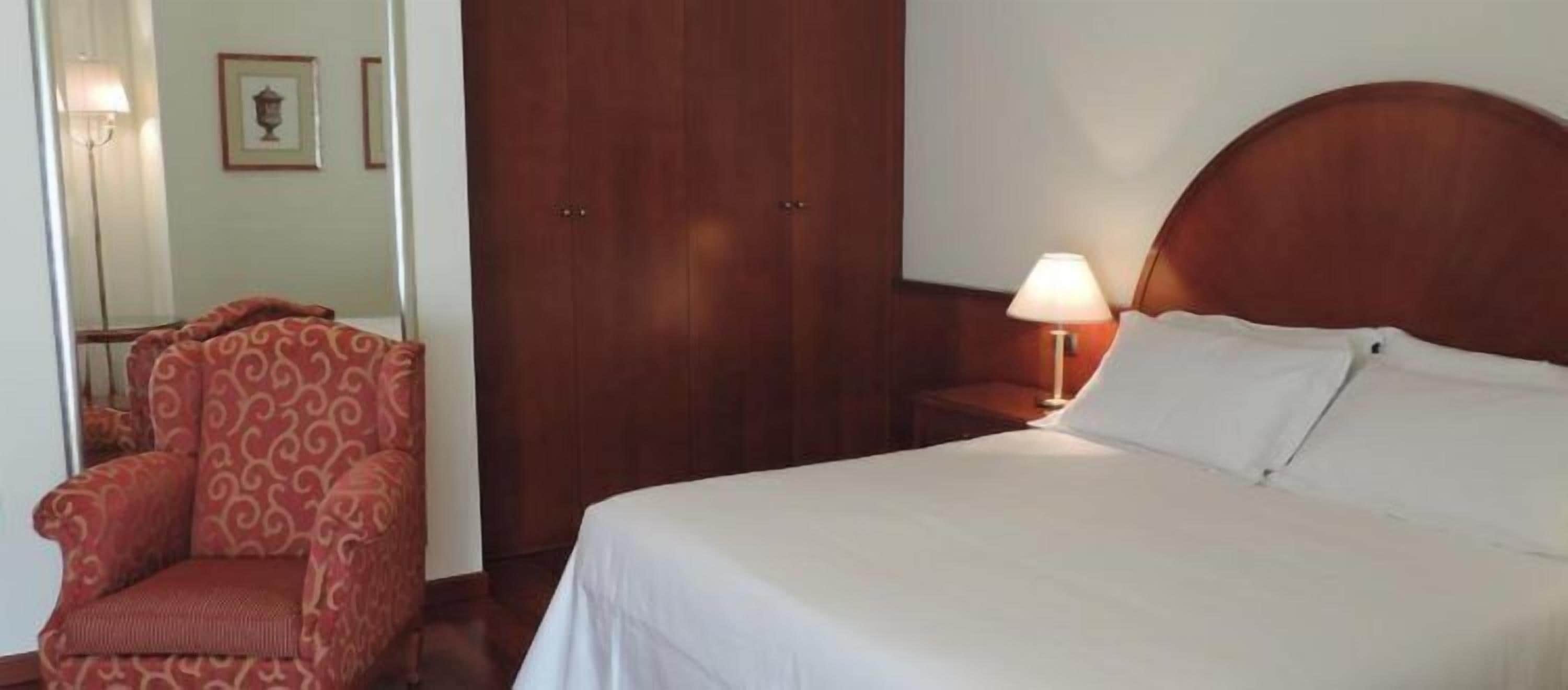 Una camera d'albergo con un letto, una sedia, un armadio e un comodino con una lampada.