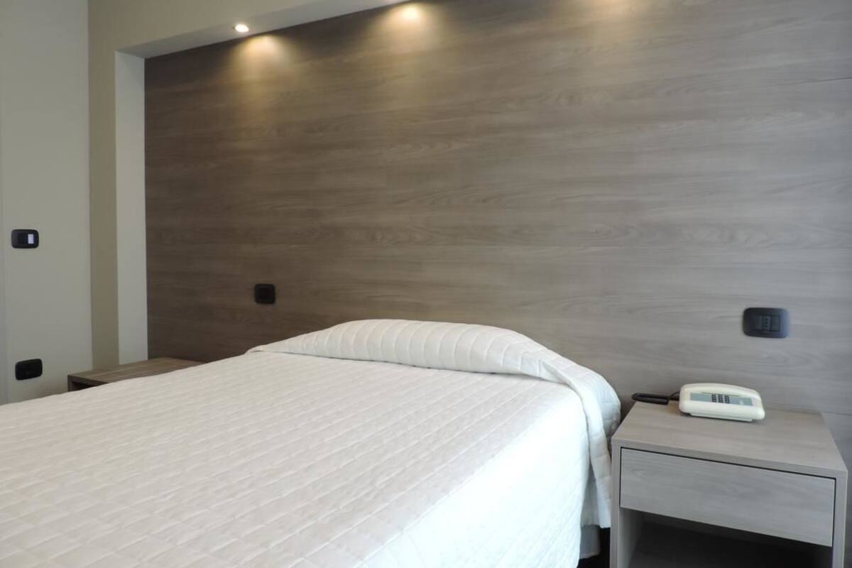 A modern hotel room with a large bed, a bedside table with a phone, and a wall-mounted light fixture.