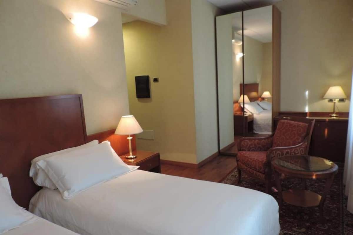 A hotel room with two beds, a sitting area with a chair and a small table, and a mirror reflecting the room.