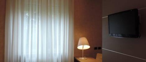 A room with a TV mounted on the wall, a lamp on a bedside table, and sheer curtains on the window.