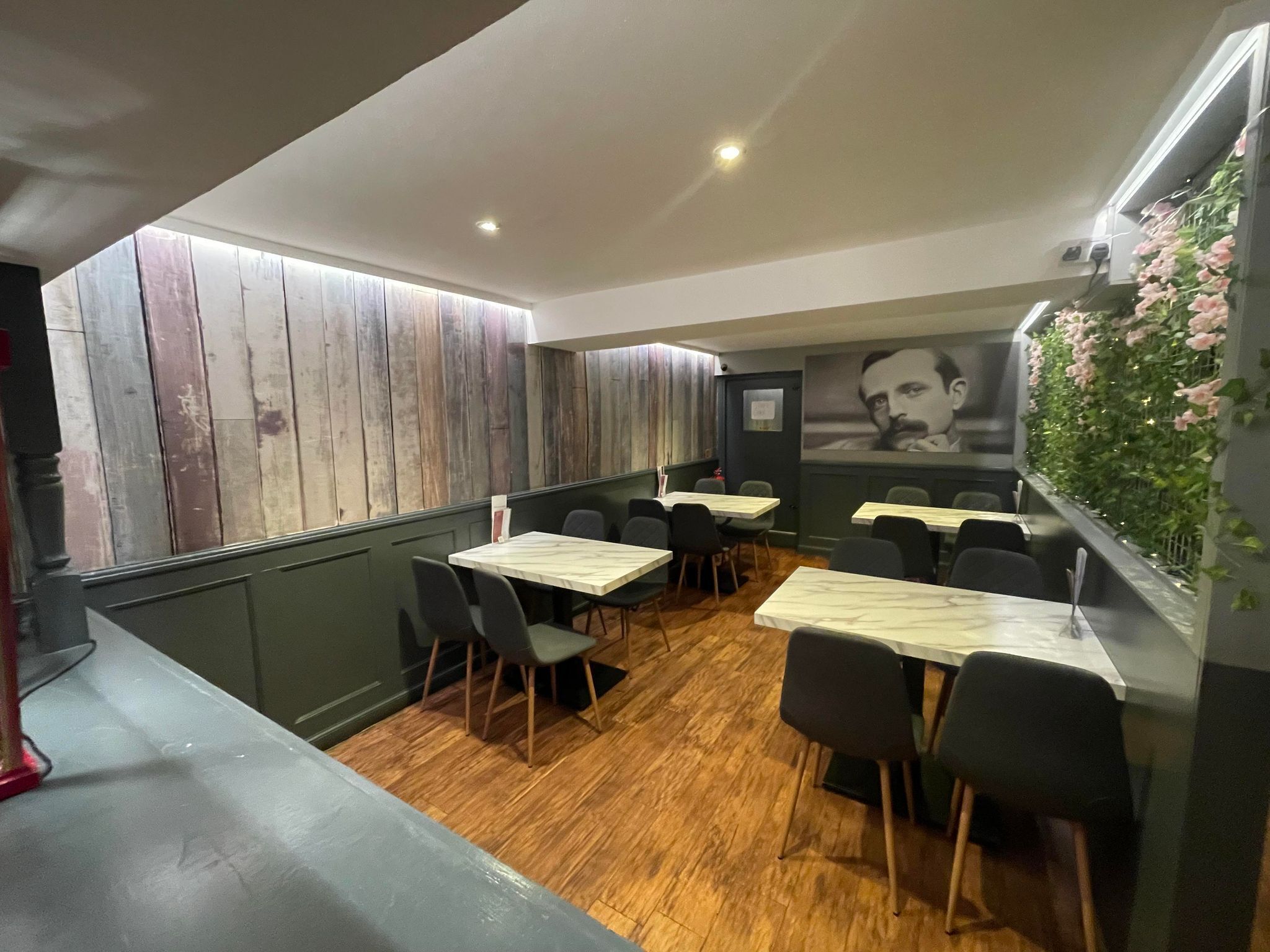 A restaurant interior with wooden panel walls, tables with chairs, and a green wall with flowers.