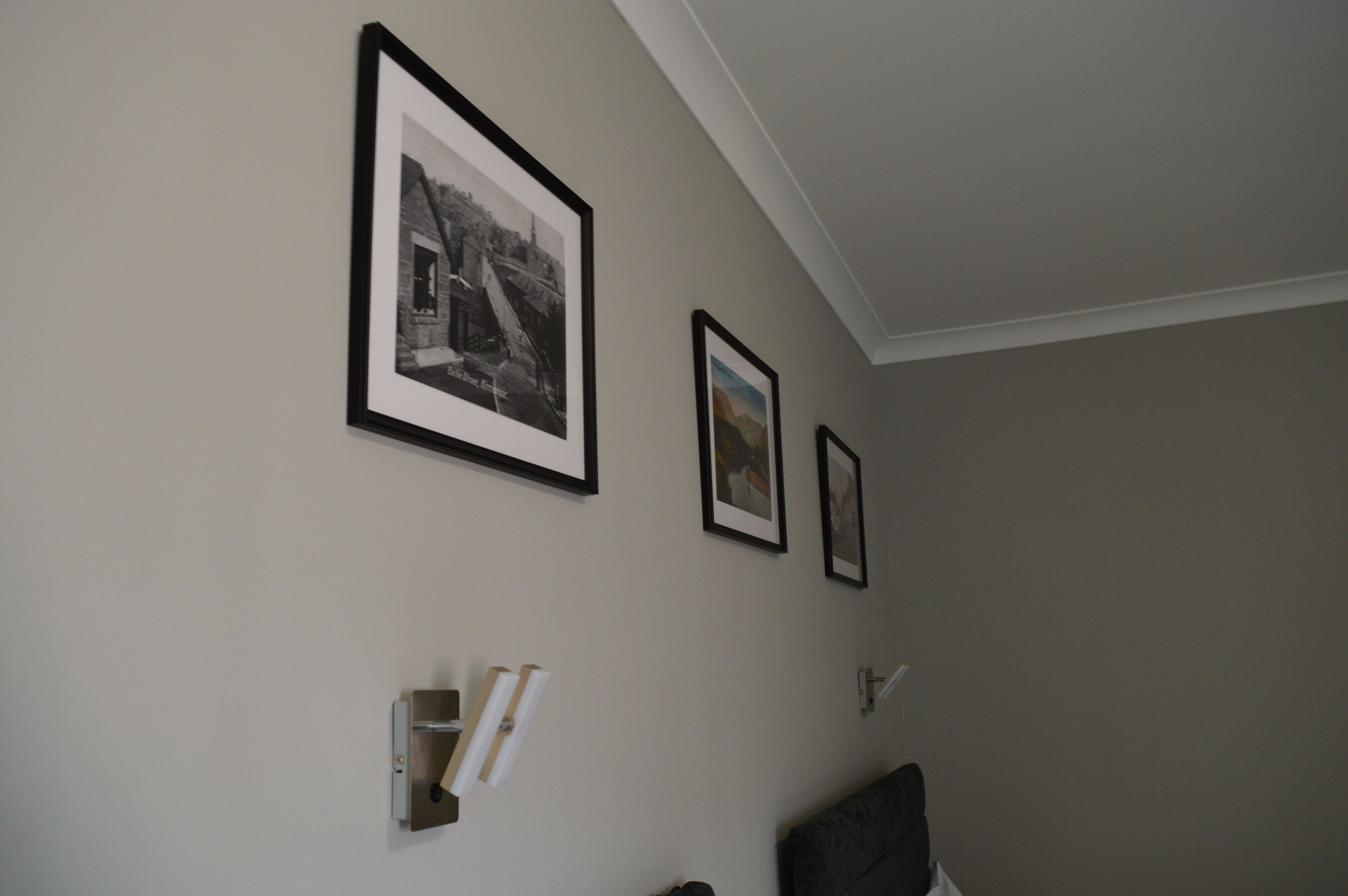 Ein Zimmer mit schlichter Wandgestaltung, drei gerahmten Bildern, einem kleinen Regal mit Büchern und einem dunkel gefärbten Stuhl.