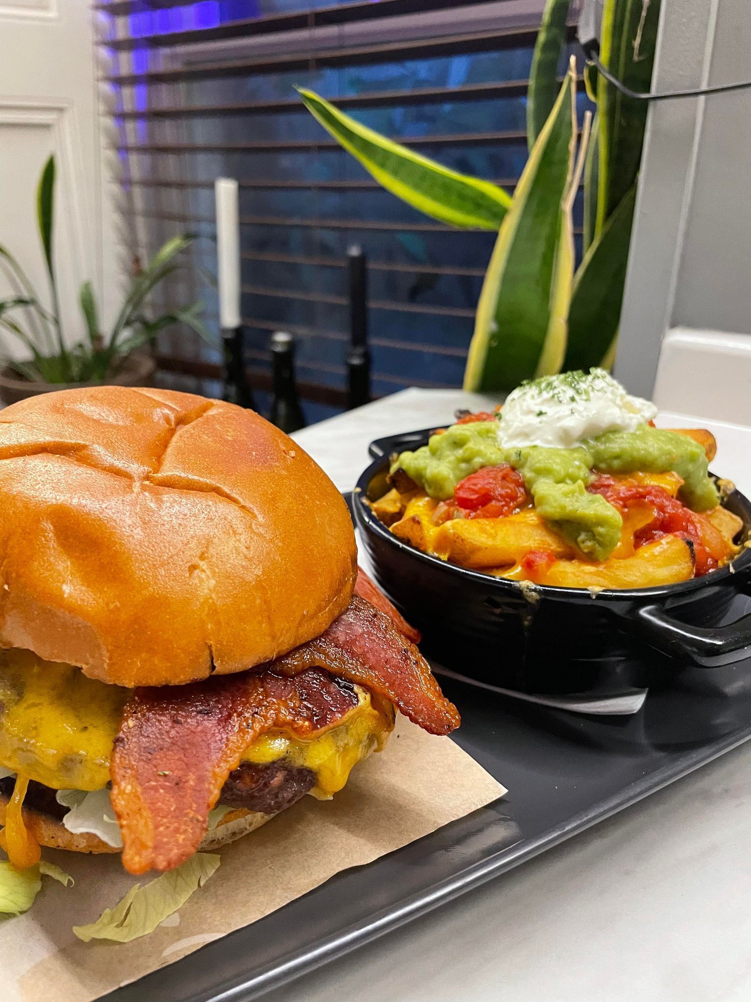 A sandwich with bacon and cheese, a bowl of nachos with guacamole and sour cream, and a plant in the background.