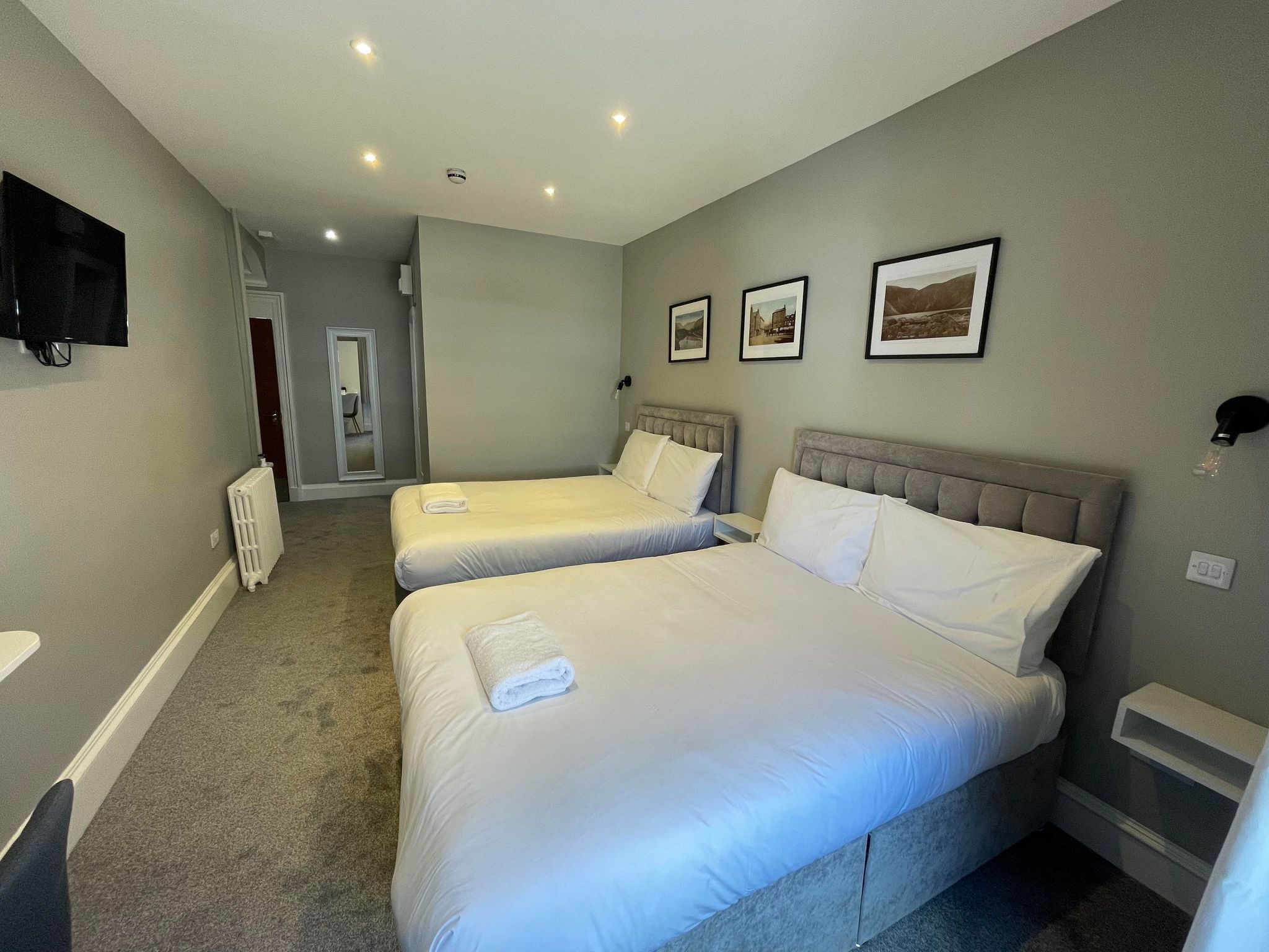 A hotel room with two beds, a flat-screen TV, a radiator, and framed pictures on the wall.