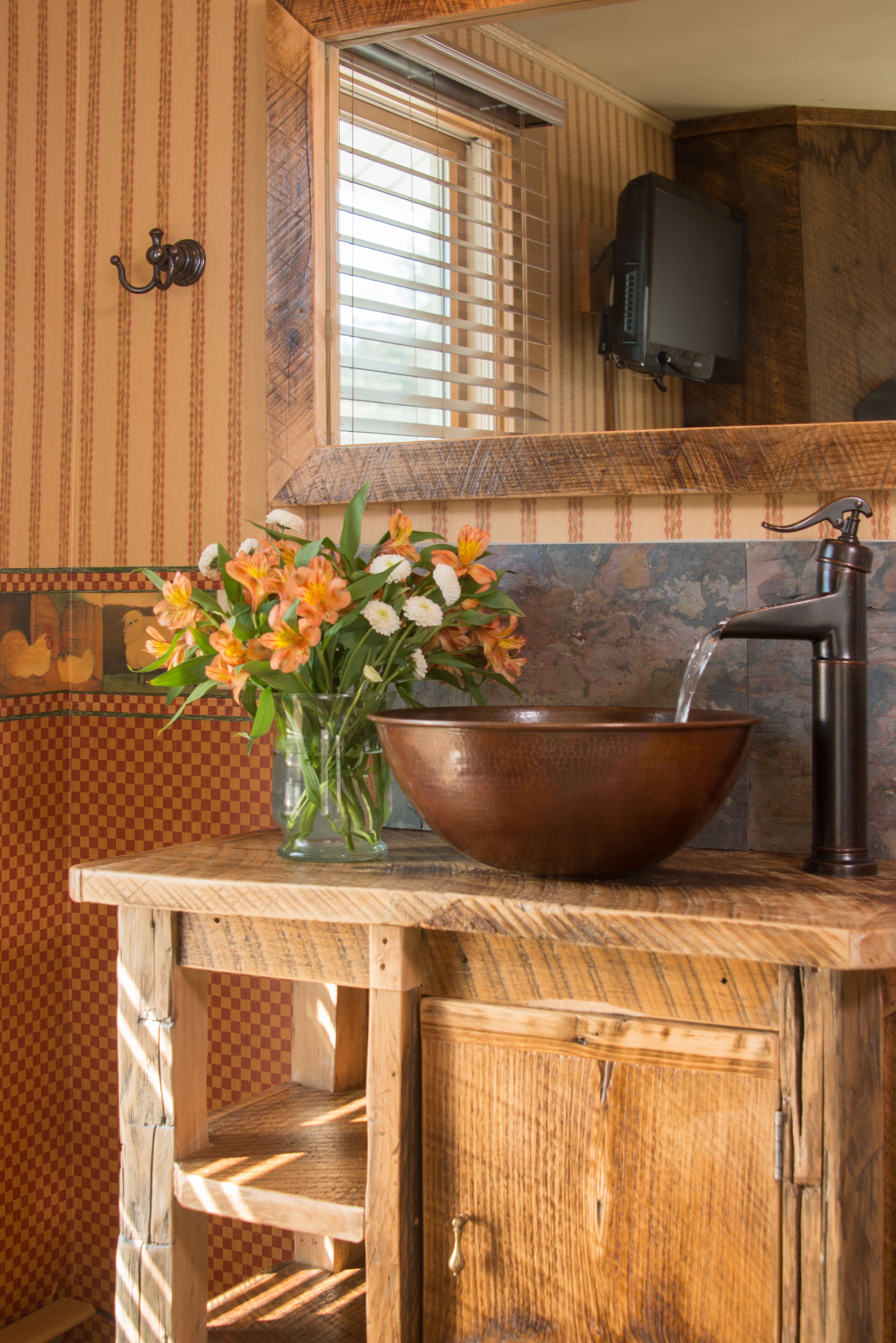 A wooden vanity with a copper sink, a vase of flowers, and a mirror above the sink.