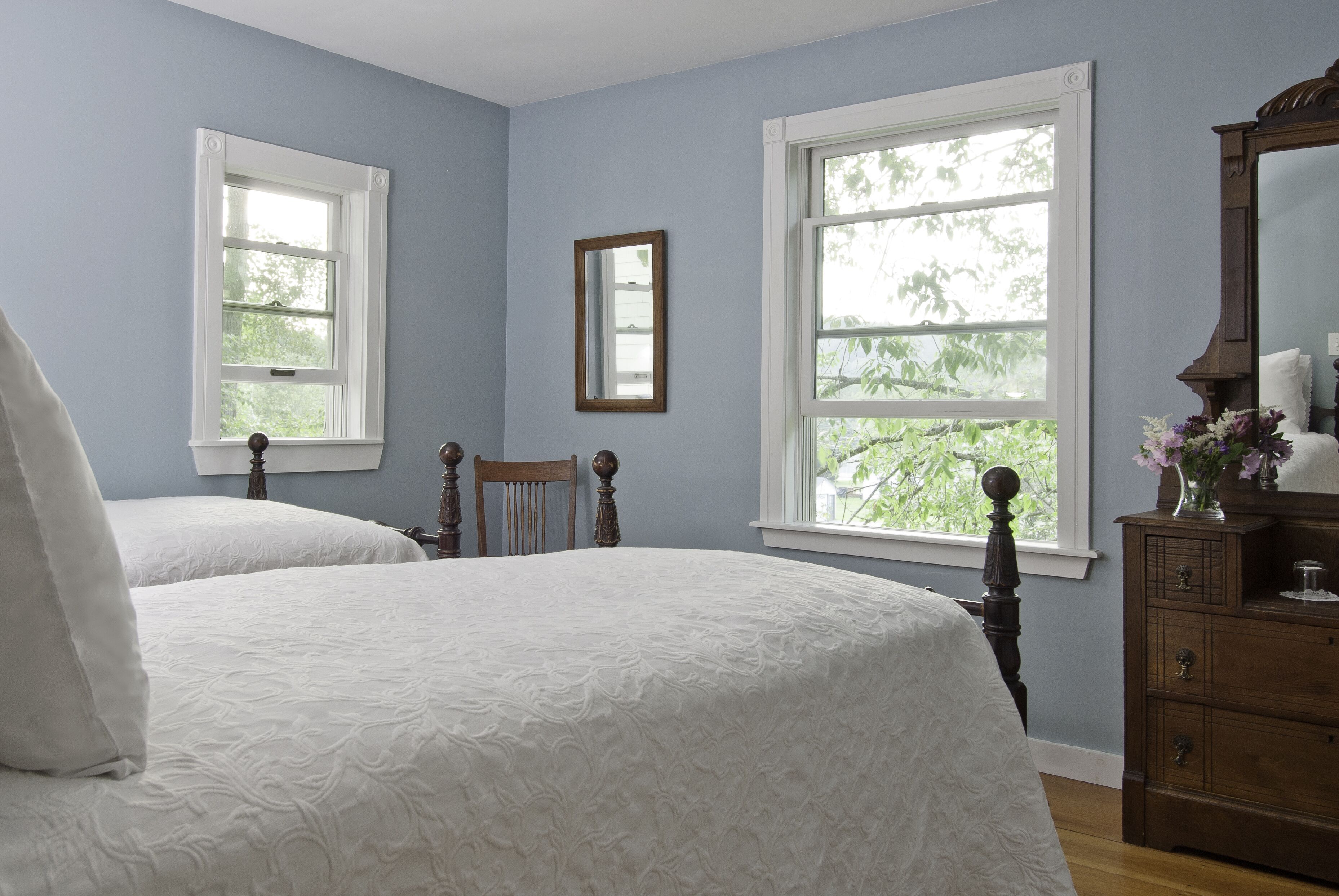 A bedroom with two single beds, a dresser, a mirror, and two windows with white frames.