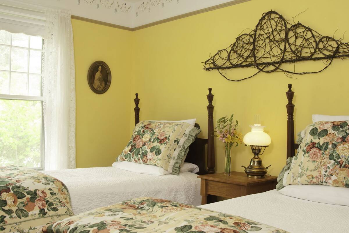 Ein Schlafzimmer mit zwei Betten, floralem Bettzeug, einem Nachttisch mit Lampe, einem Wandbild und einem Fenster mit Vorhängen.
