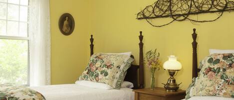 A bedroom with two beds, floral bedding, a bedside table with a lamp, a wall-mounted picture, and a window with curtains.