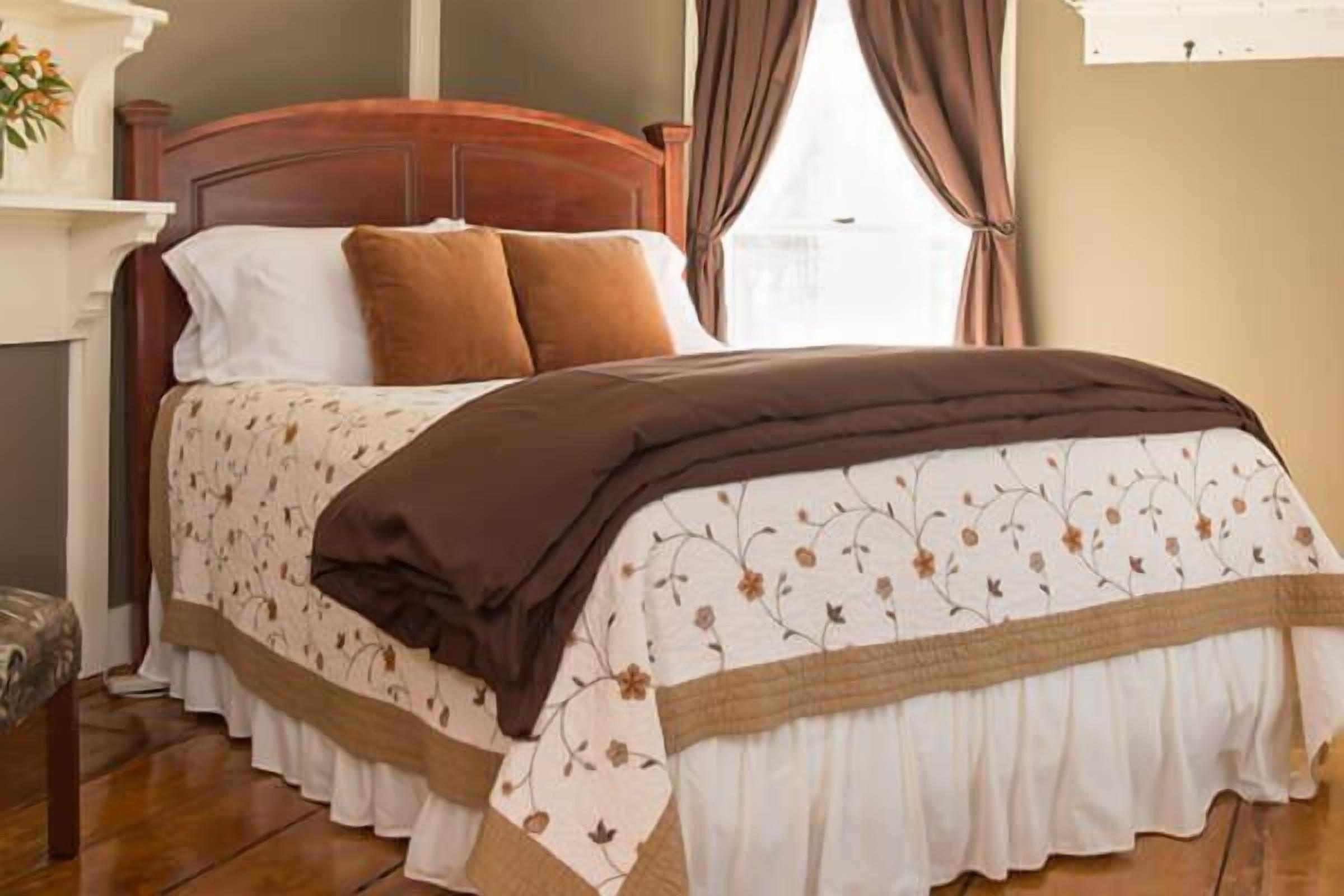 A neatly made bed with a floral-patterned bedspread and brown pillows, a wooden headboard, and a window with curtains.