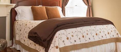 A neatly made bed with a floral-patterned bedspread and brown pillows, a wooden headboard, and a window with curtains.