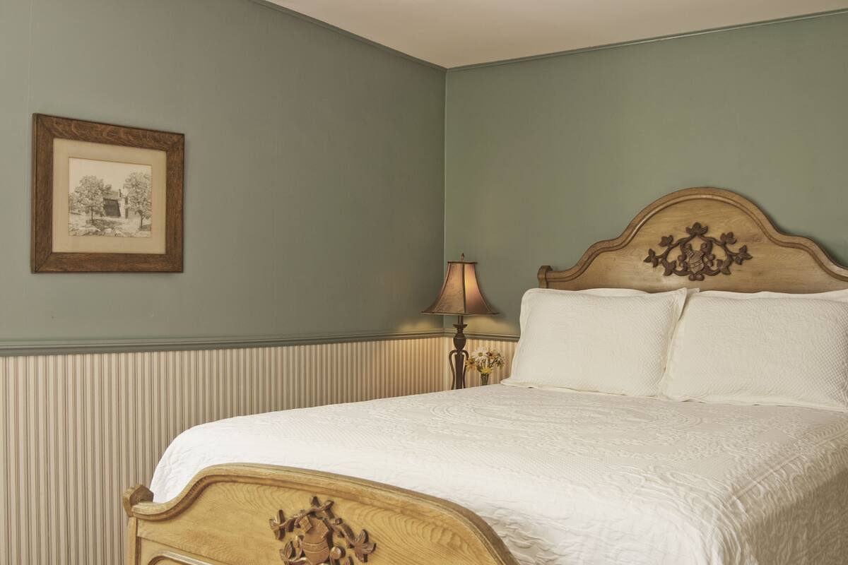 Ein ordentlich bezogenes Bett mit Holz-Kopfteil, ein gerahmtes Bild an der Wand und eine Lampe neben dem Bett.