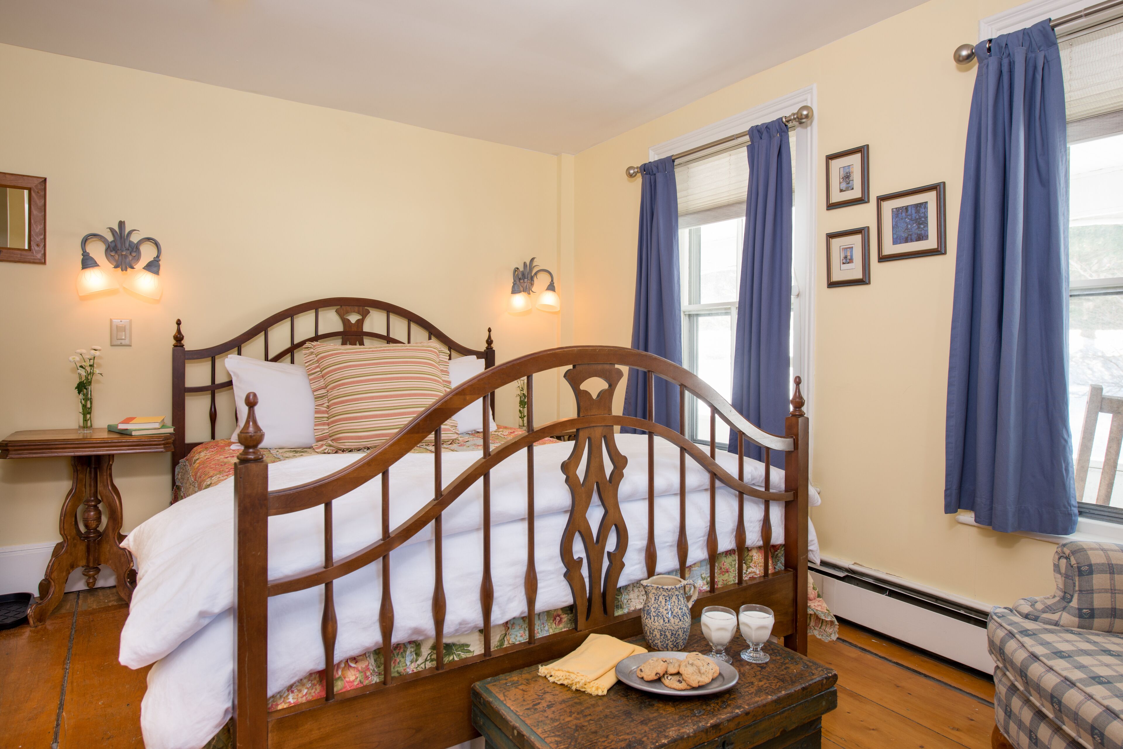 Un dormitorio con una cama de madera, una mesita de noche con lámpara, una ventana con cortinas y una pequeña mesa con un plato de galletas.