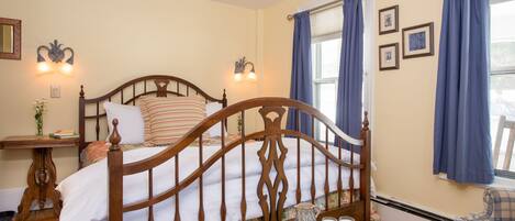 Un dormitorio con una cama de madera, una mesita de noche con lámpara, una ventana con cortinas y una pequeña mesa con un plato de galletas.