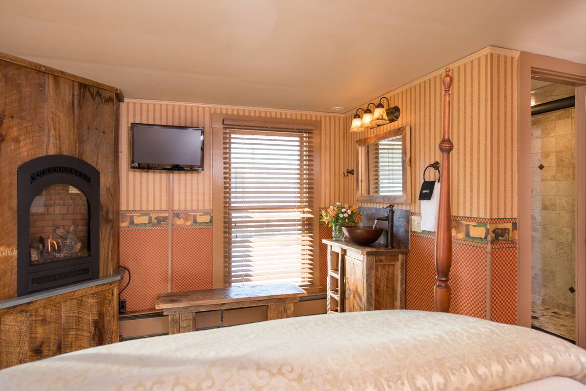 Ein Schlafzimmer mit Kamin, Fernseher, einer hölzernen Bank sowie einem Badezimmer inklusive Spiegel und Handtuchhalter.