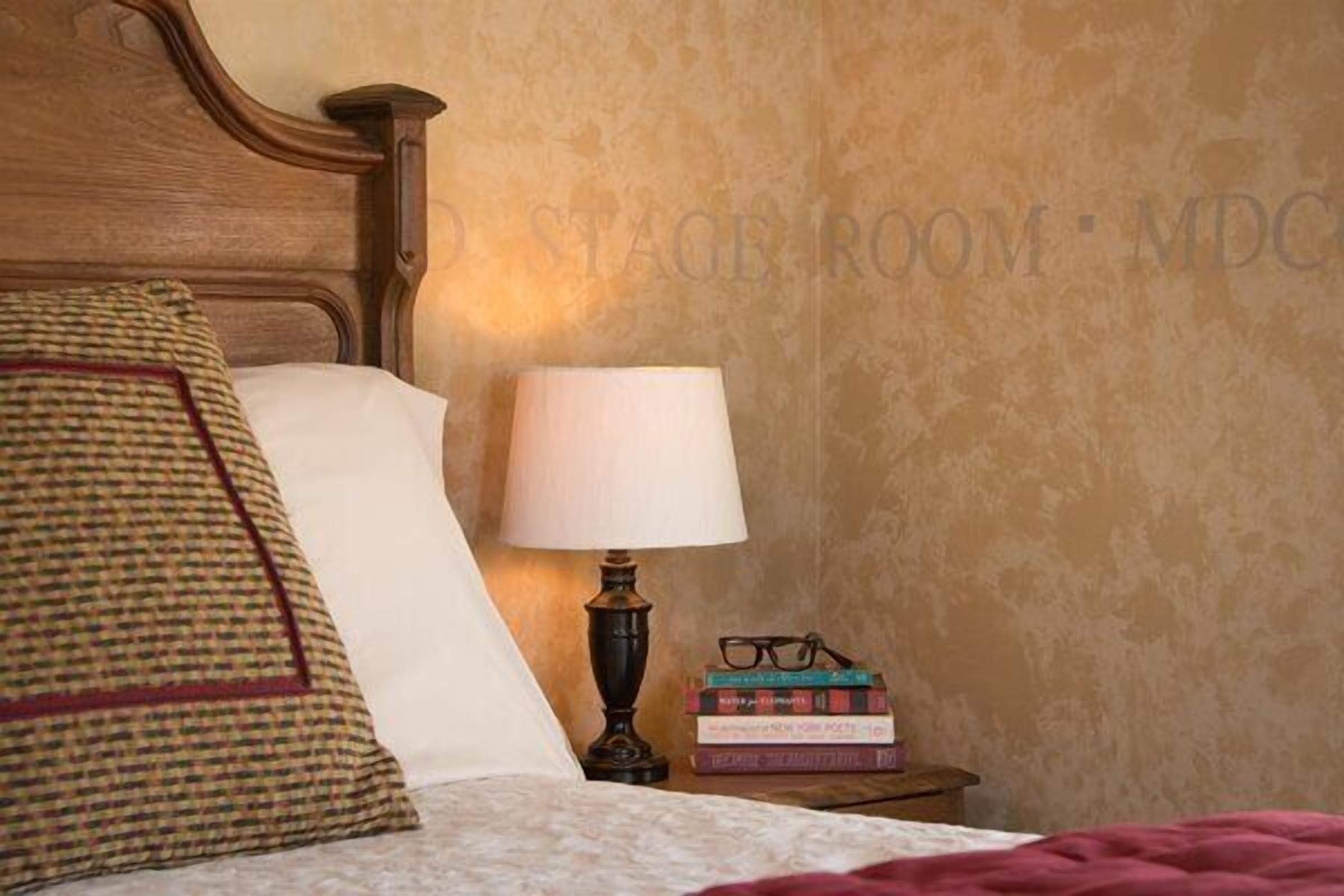 A neatly made bed with a checkered pillow, a white pillow, and a red blanket. There is a bedside table with a lamp and a stack of books. The wall has a textured pattern.