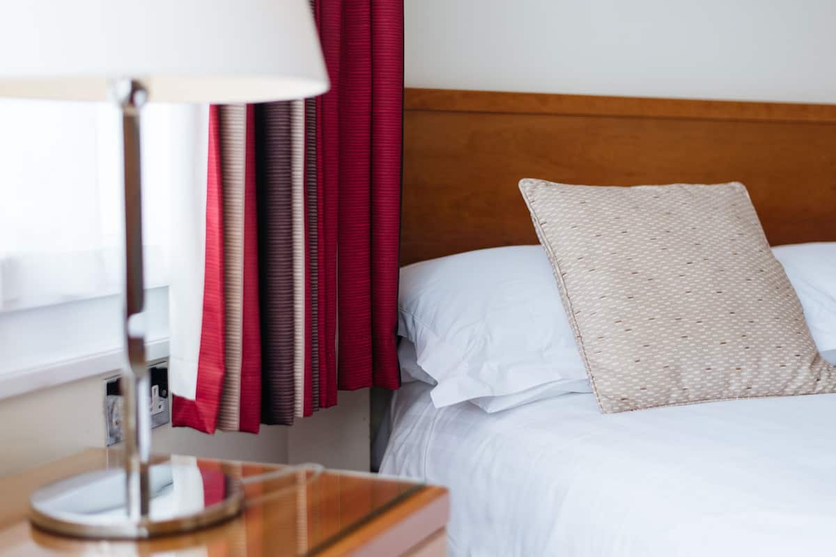 A neatly made bed with a patterned pillow, a bedside lamp, and a window with red curtains.