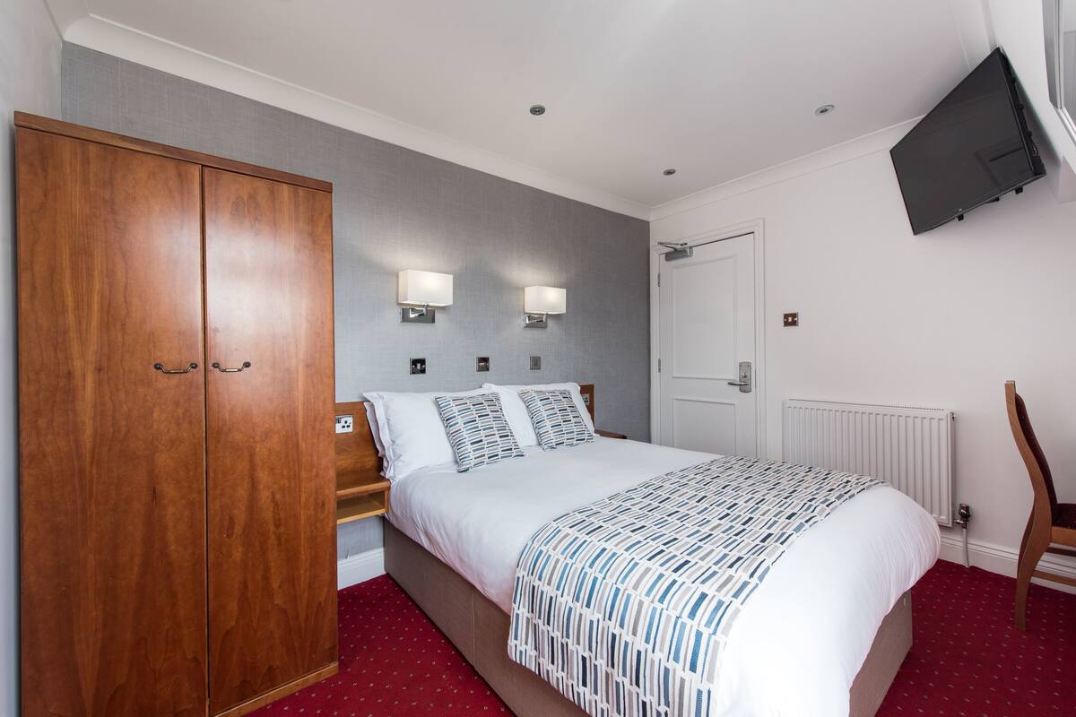 A bedroom with a wooden wardrobe, a bed with a patterned bedspread, bedside lamps, a flat-screen TV mounted on the wall, and a radiator.